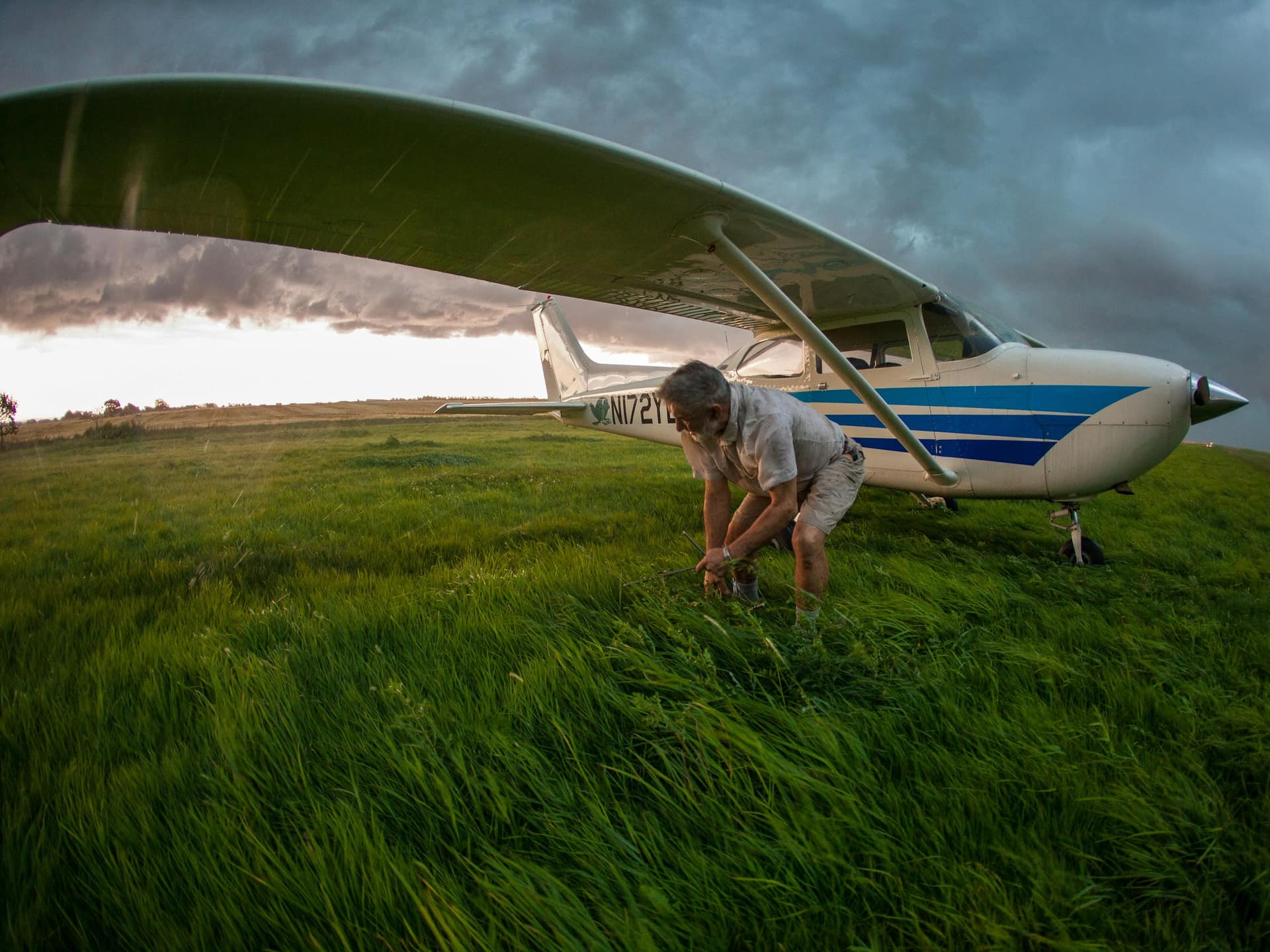 A pilot kneeling in tall grass beside a blue-striped high-wing airplane under a stormy sky